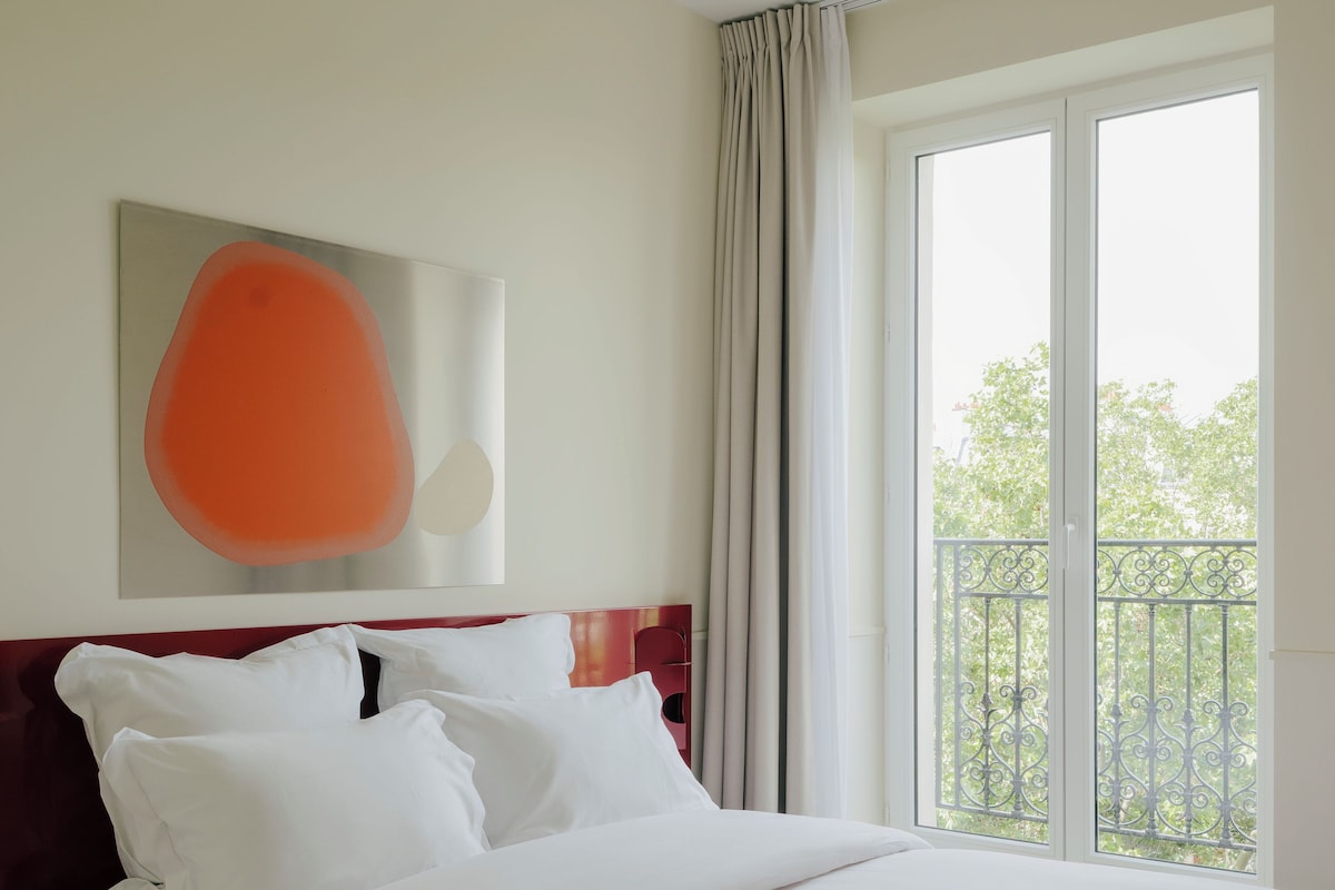 A hotel room with a bed, a large window with a view, a ceiling light, and a wall-mounted artwork.