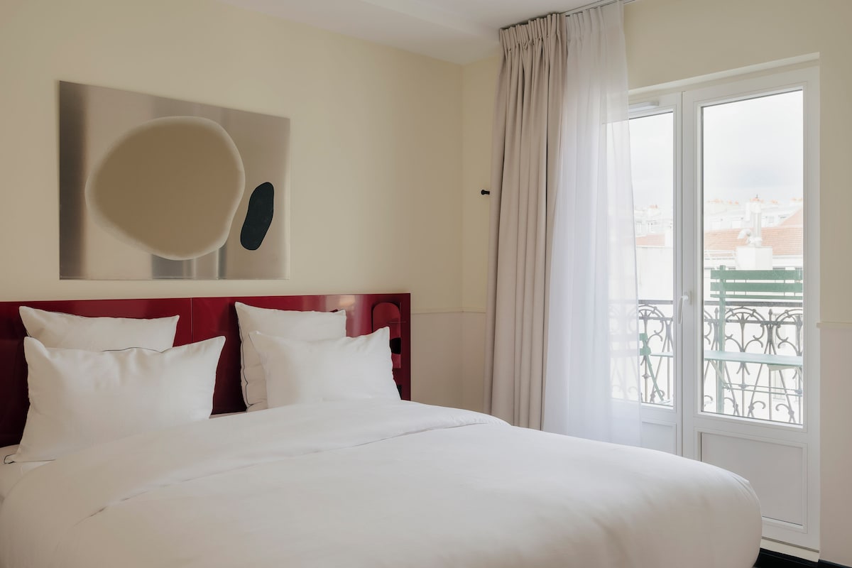 A hotel room with a large bed, a view of a balcony with a railing, and a painting on the wall.