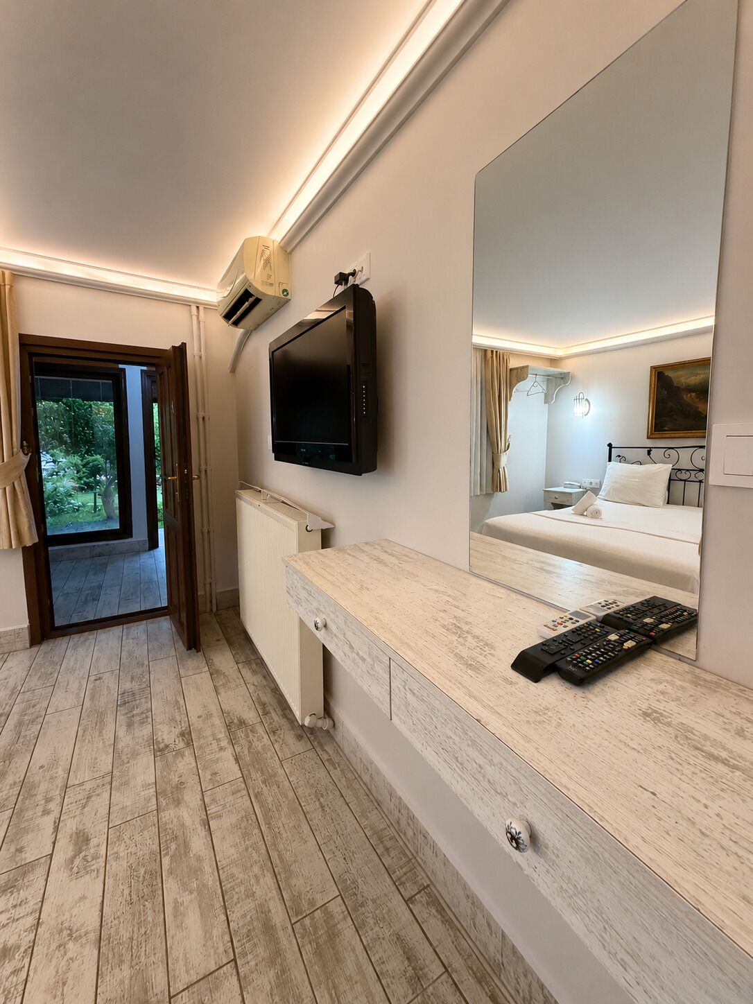 A hotel room with a TV mounted on the wall, a bed with a headboard, a bedside table with a remote control, and a view of greenery outside.