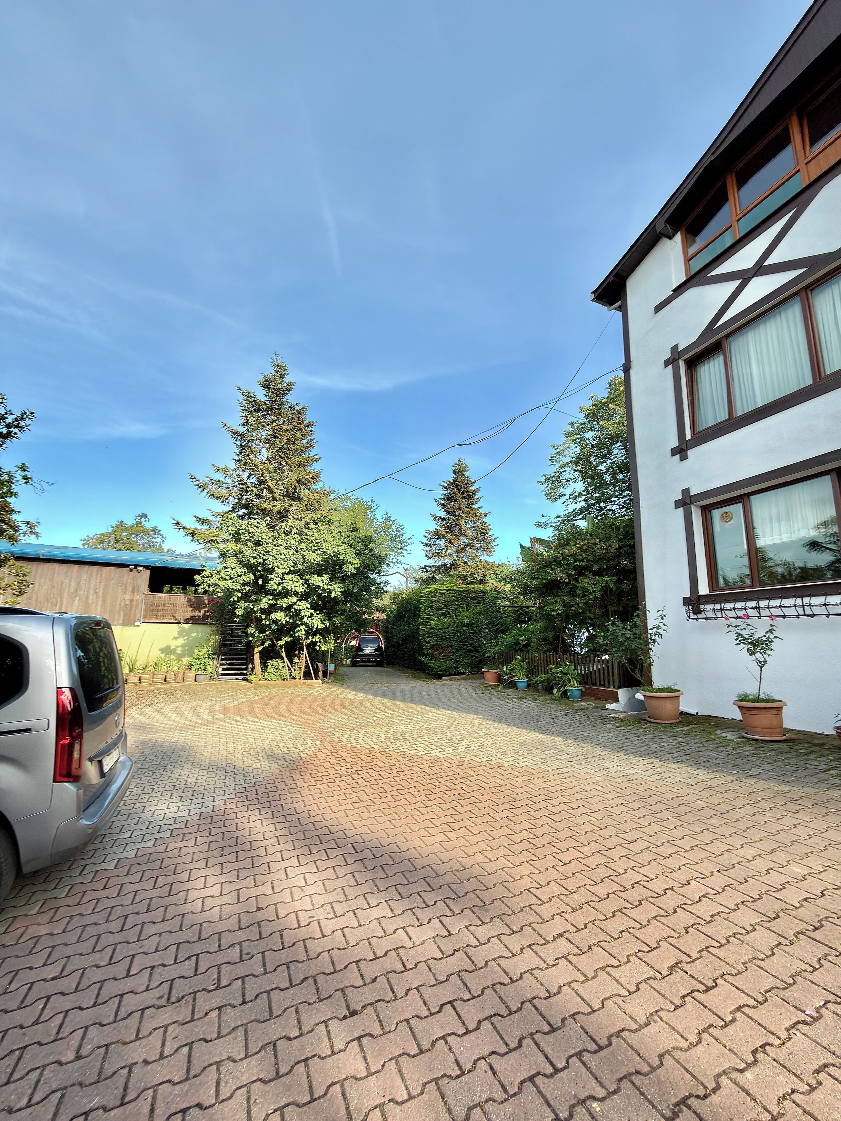A paved driveway leading to a building with a white exterior and dark trim. There are potted plants along the path and a car parked on the left.