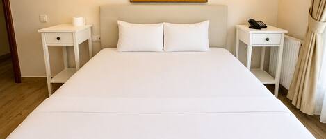 A neatly made bed with white linens, two bedside tables with lamps, a painting above the bed, and a radiator on the wall.