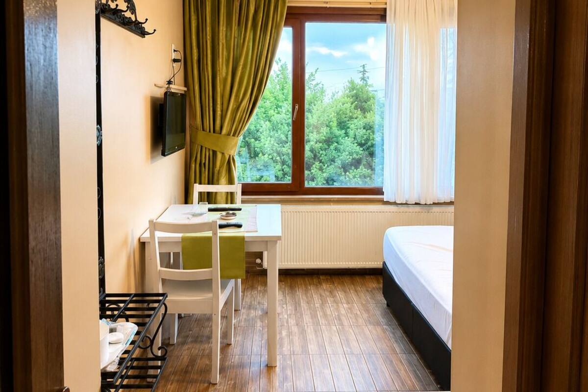 Una habitación pequeña con una cama, una mesa con sillas, un radiador, una ventana con cortinas y un televisor en la pared.