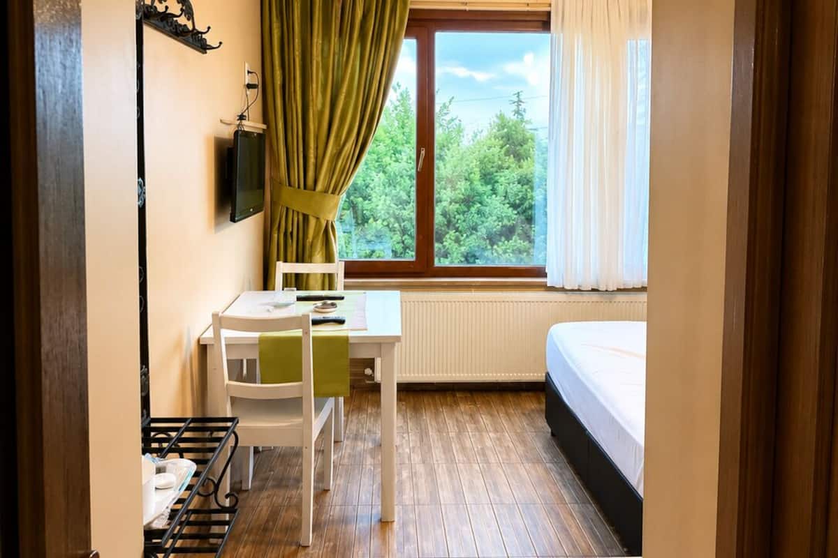 Une petite pièce comprenant un lit, une table avec des chaises, un radiateur, une fenêtre avec des rideaux et un téléviseur fixé au mur.