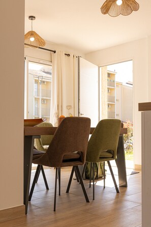 A dining area with a wooden table and chairs, a hanging light fixture, and a view of buildings through an open door.