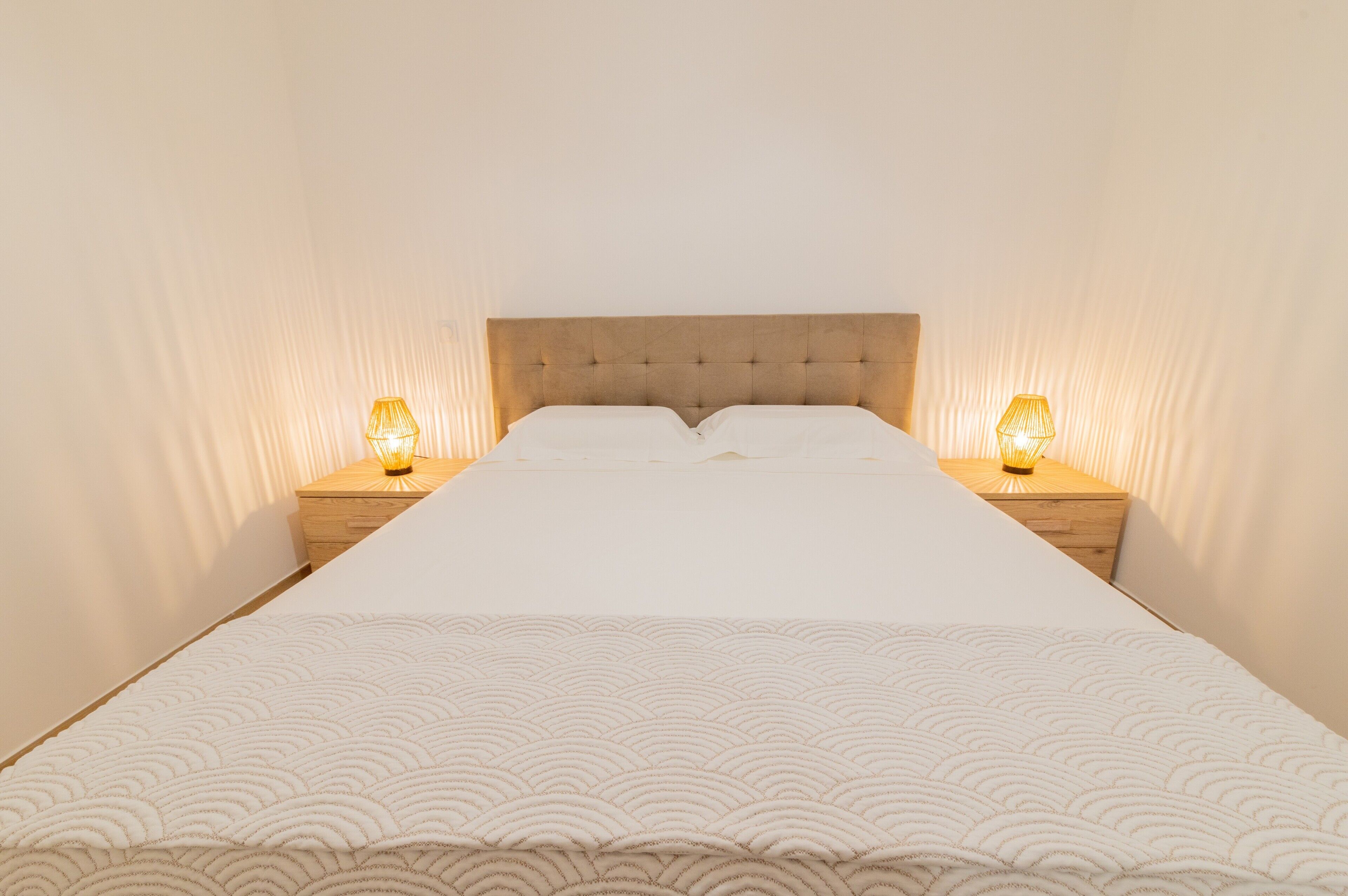 A neatly made bed with a tufted headboard, two bedside lamps, and a plain wall.