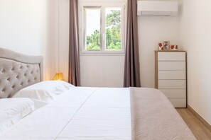 A neatly made bed with a headboard, a bedside lamp, a window with curtains, a dresser, and an air conditioning unit on the wall. - Apartment 'Residence La Dolce Vita' with Sea View, Wi-Fi and Air Conditioning (Borgo)