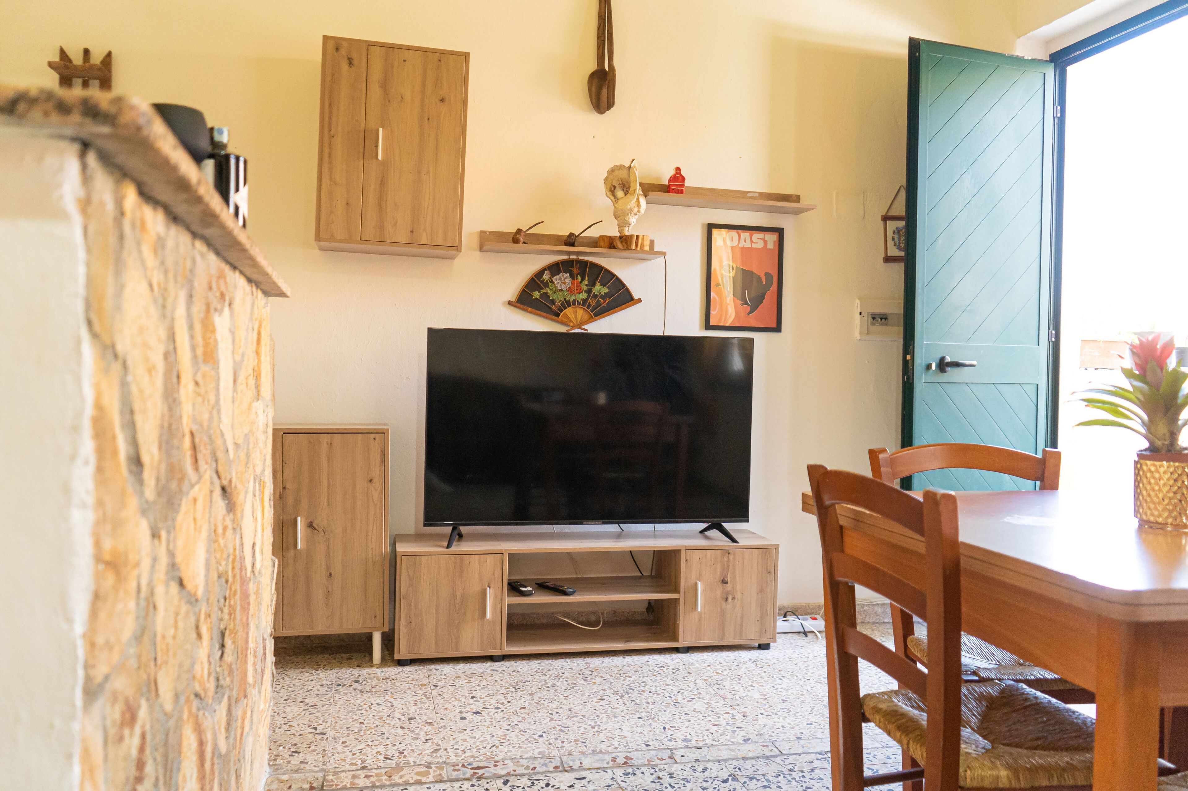 A living room with a television, wooden cabinets, a dining table with chairs, and a door leading outside.