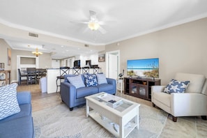 Ein Wohnzimmer mit Sofa, Sessel, Couchtisch und Fernseher. Im Hintergrund ist eine Küche zu sehen.