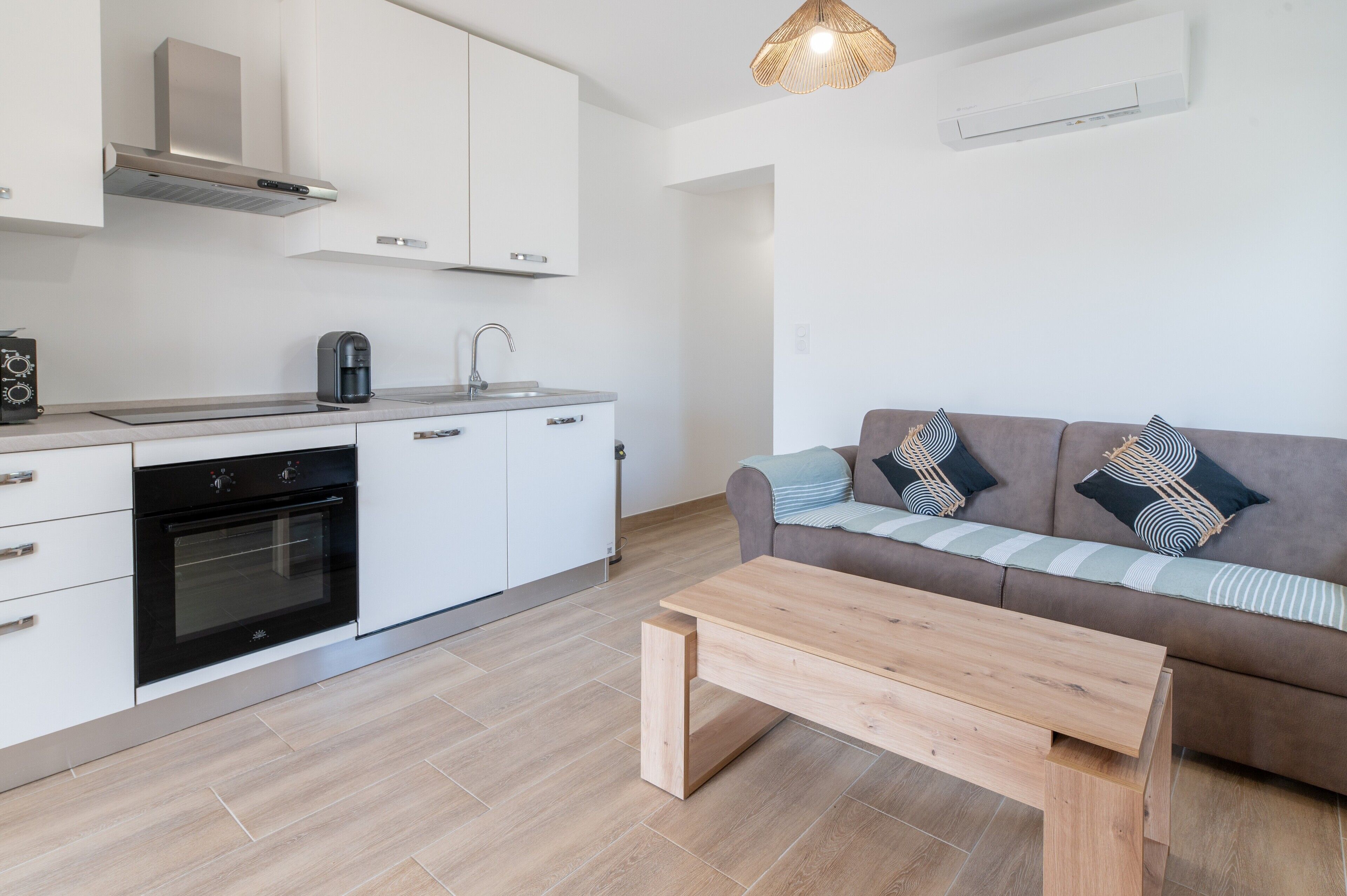 Eine moderne Küche mit weißen Schränken, einem eingebauten Ofen und einer Kaffeemaschine. Im Wohnbereich befindet sich eine Holzbänke mit einem braunen Sofa und gemusterten Kissen.