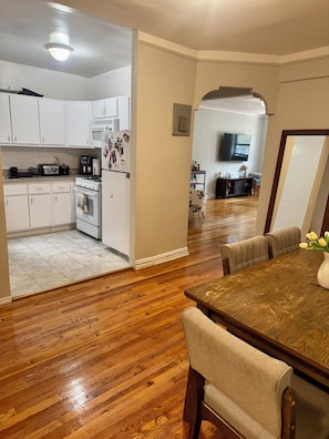 A kitchen with white cabinets, a stove, and a refrigerator. There is a dining area with a wooden table and chairs. - Large 2-bedroom apartment in Sunnyside, 2 block from subway (Sunnyside)