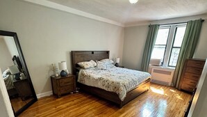 Ein Schlafzimmer mit Holzboden, einem Bett mit Kopfteil, einem Nachttisch, einer Kommode, einem Fenster mit Vorhängen und einer Deckenleuchte.
