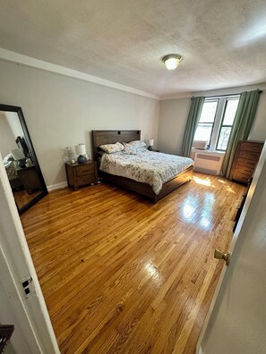 A bedroom with a wooden floor, a bed with a headboard, a nightstand, a dresser, a window with curtains, and a ceiling light. - Large 2-bedroom apartment in Sunnyside, 2 block from subway (Sunnyside)