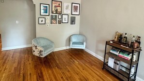 Ein Zimmer mit Holzboden, zwei hellblaue Stühle, ein Bücherregal mit Büchern und Flaschen sowie gerahmte Bilder an der Wand.