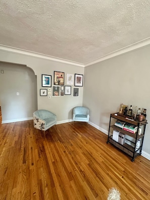 A room with wooden flooring, two light blue chairs, a bookshelf with books and bottles, and framed pictures on the wall. - Large 2-bedroom apartment in Sunnyside, 2 block from subway (Sunnyside)
