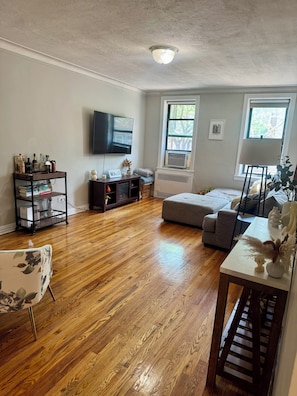 A living room with hardwood flooring, a grey sofa, a TV mounted on the wall, and a window with an air conditioning unit. - Large 2-bedroom apartment in Sunnyside, 2 block from subway (Sunnyside)