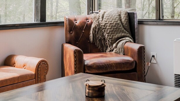 Une pièce avec une grande fenêtre, un fauteuil en cuir, une table en bois et une petite commode en bois.