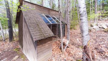 A wooden birdhouse with a sloped roof and a window, surrounded by trees and forest floor.