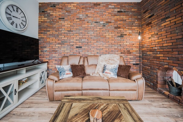 Un salón con pared de ladrillo, sofá de cuero, mesa de centro de madera y un televisor sobre un mueble blanco.