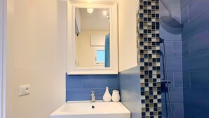 Un baño con lavamanos blanco, espejo y dos cajones, ducha con azulejos azules y suelo de baldosas.
