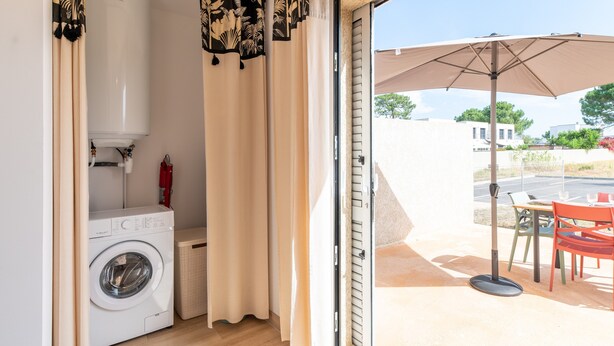 Un área de lavandería con lavadora, extintor y puerta corrediza de vidrio que da a un patio exterior con mesa, sillas y sombrilla.