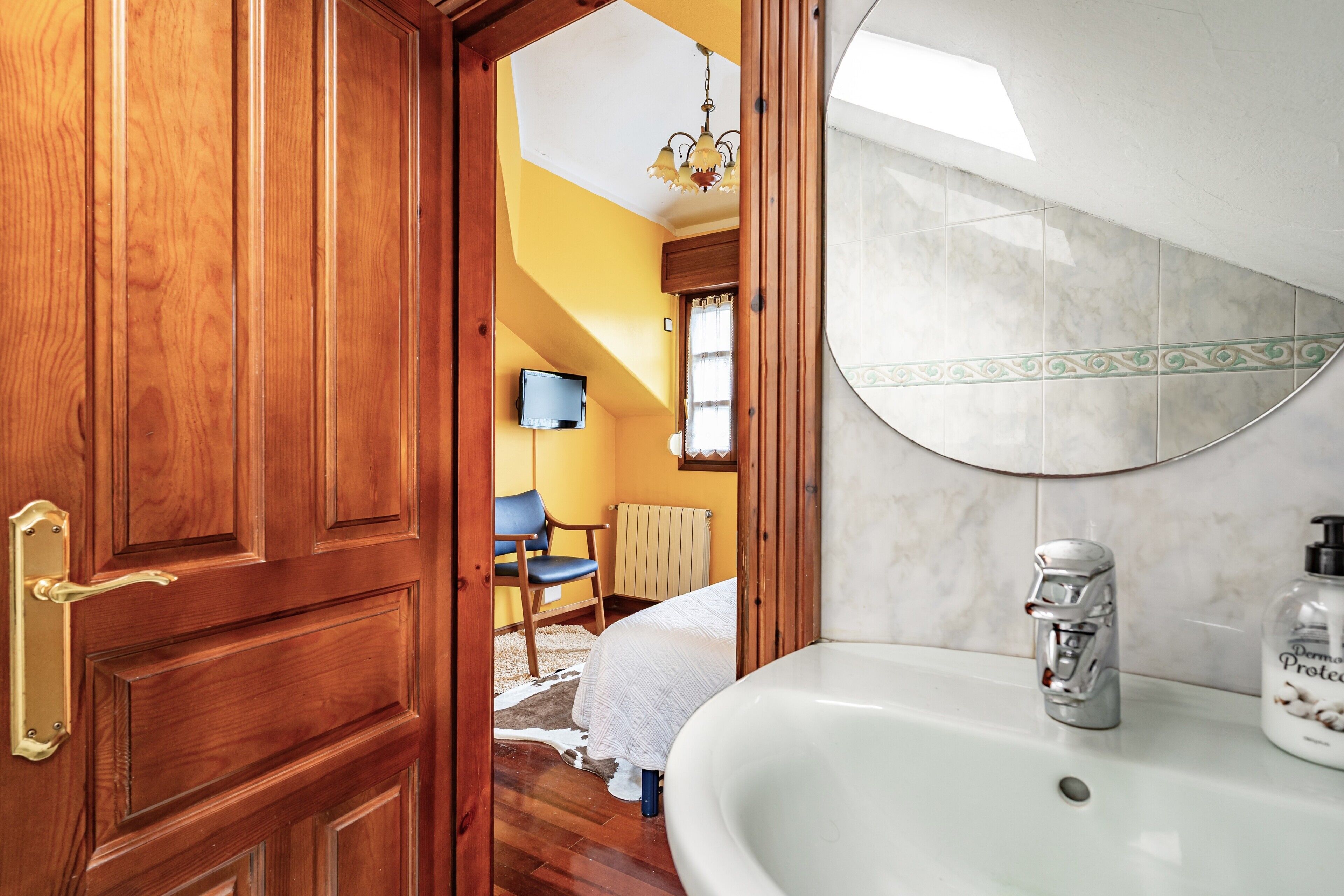 A wooden door, a white sink, a mirror, a bed, a chair, and a television in a room.