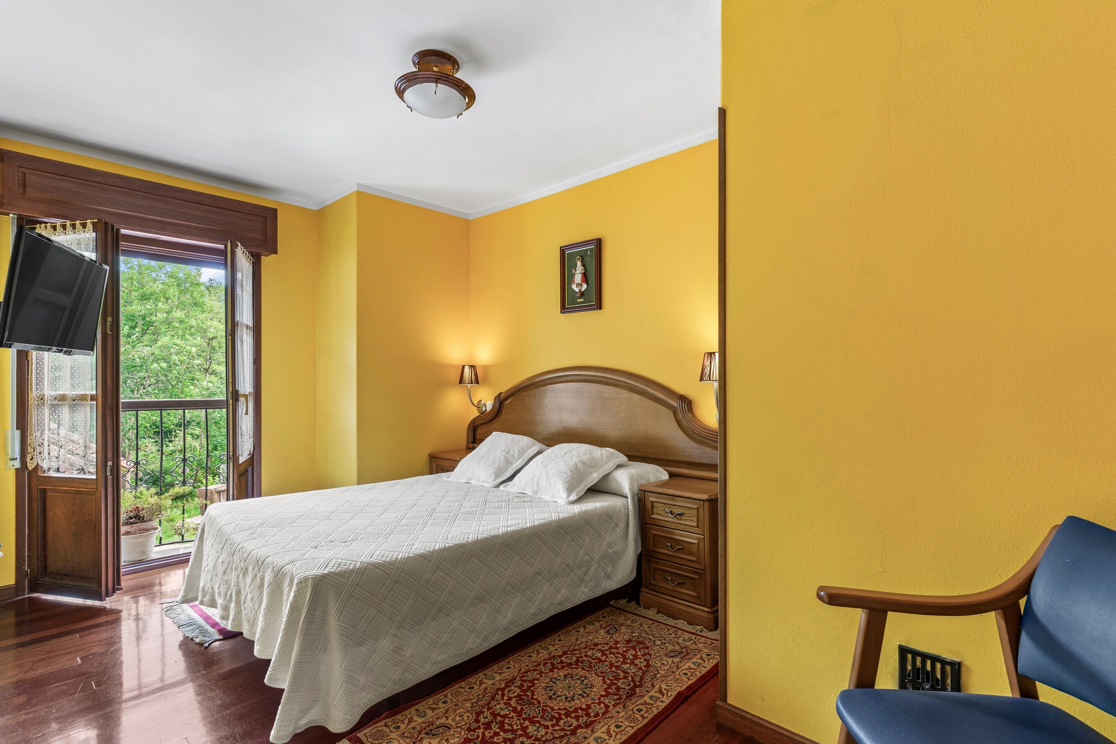 Un dormitorio con cama, mesitas de noche de madera, televisor, ventana con cortinas y puerta.
