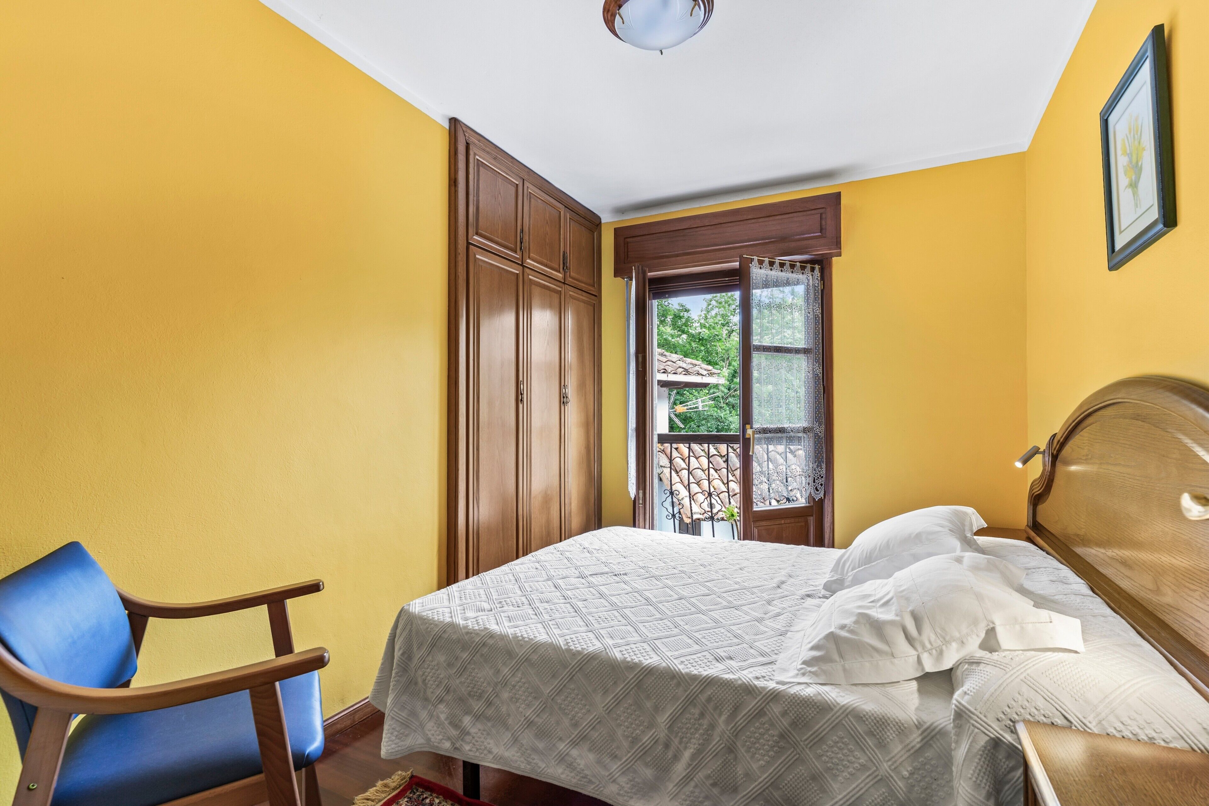 A bedroom with a bed, a chair, a wardrobe, and a window with a view of greenery.