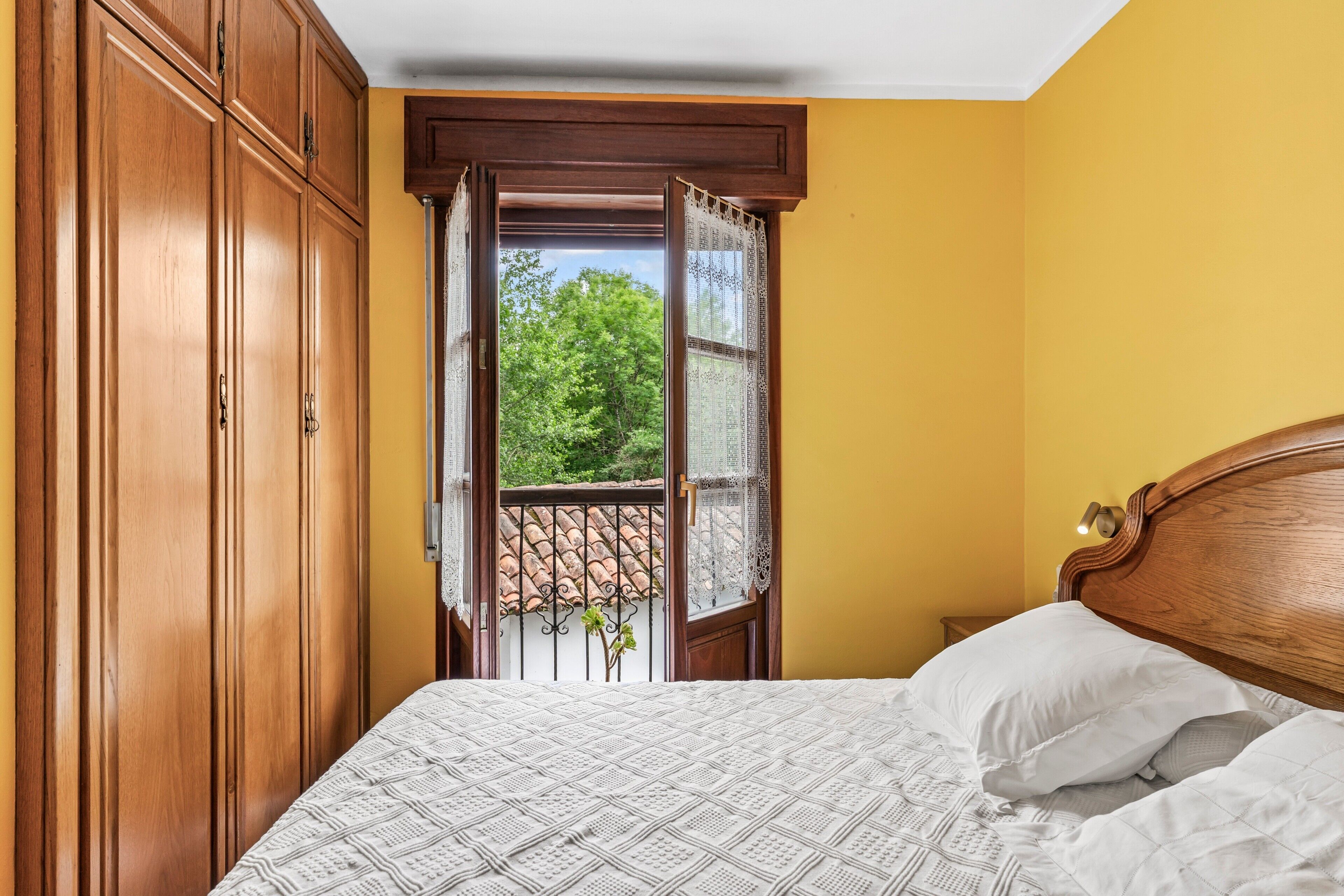 A bedroom with a wooden wardrobe, a bed with white bedding, and a view through an open window.