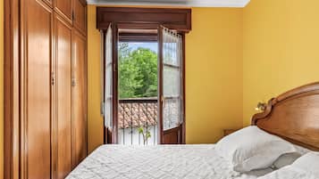A bedroom with a wooden wardrobe, a bed with white bedding, and a view through an open window.