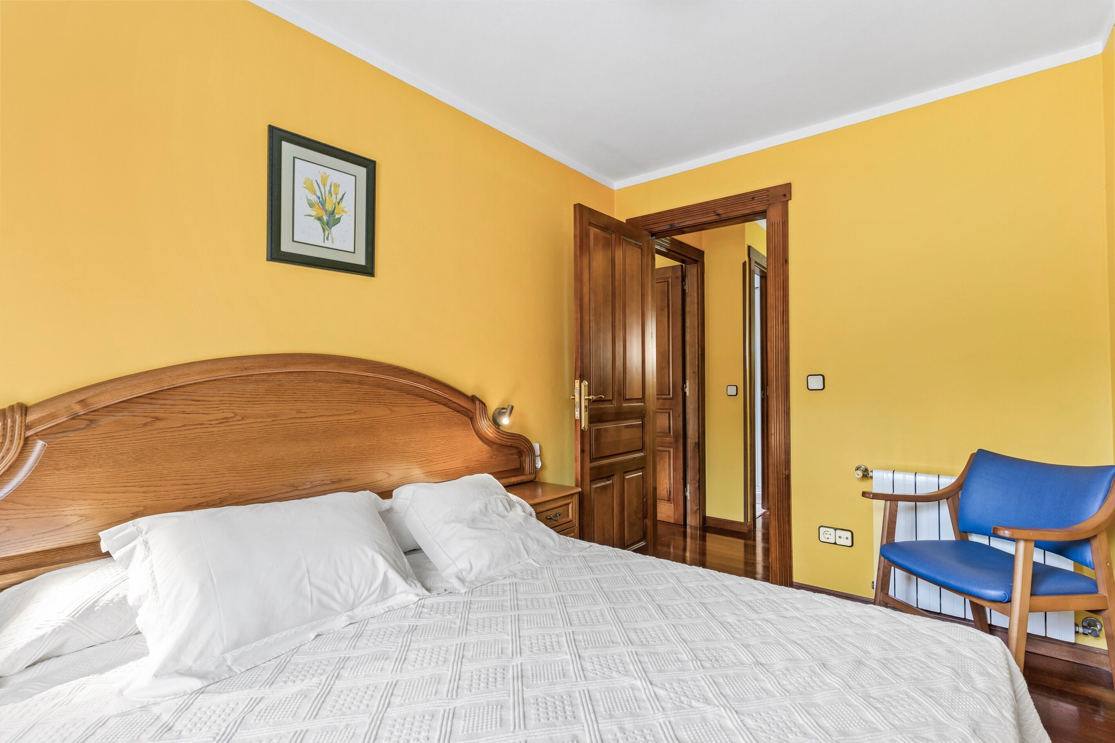 A bedroom with a wooden headboard, a bed with white bedding, a blue chair, and a wooden wardrobe.