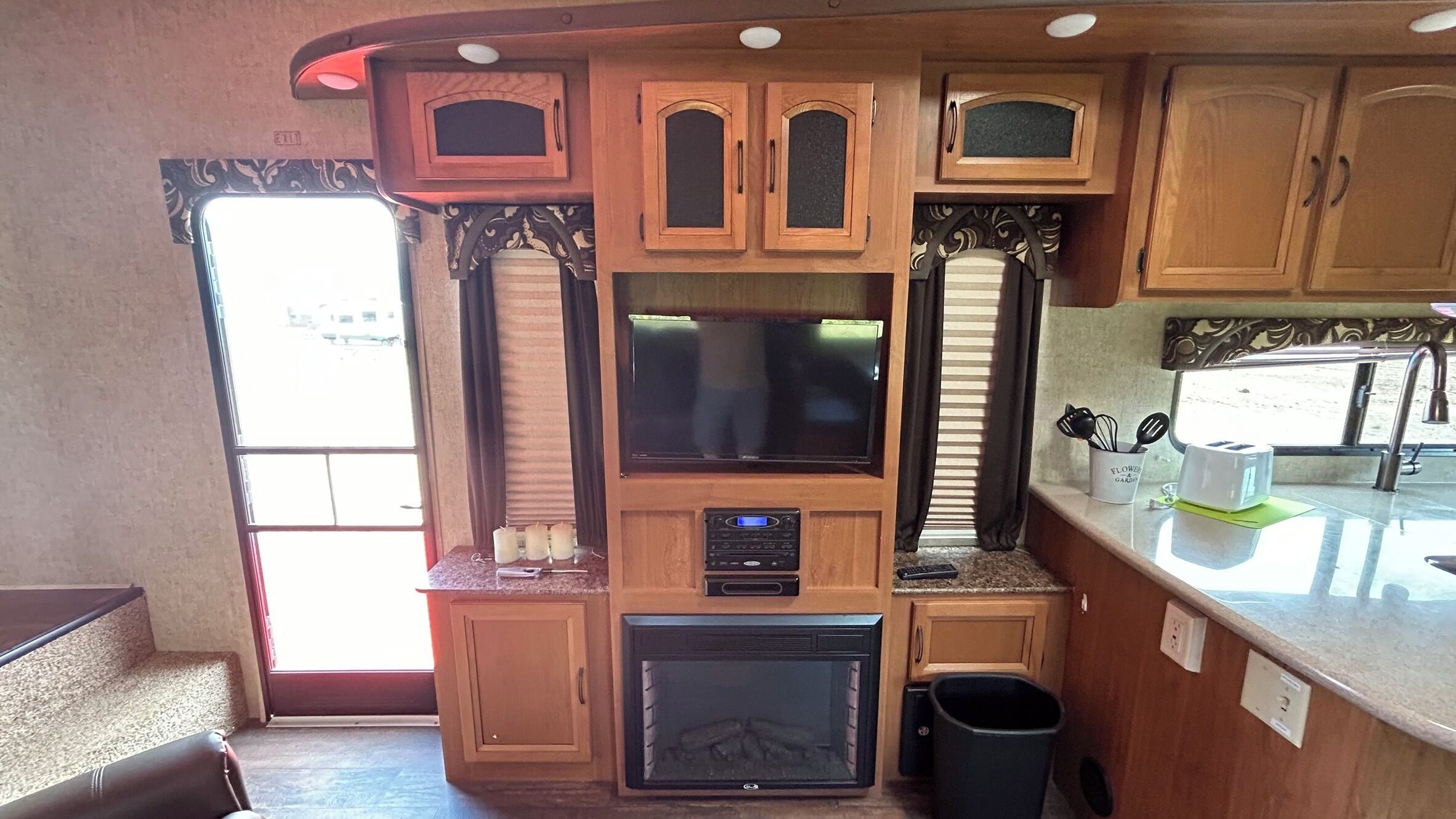 Cocina compacta para caravana con armarios de madera, microondas integrado, pequeña estufa y ventana con cortinas.