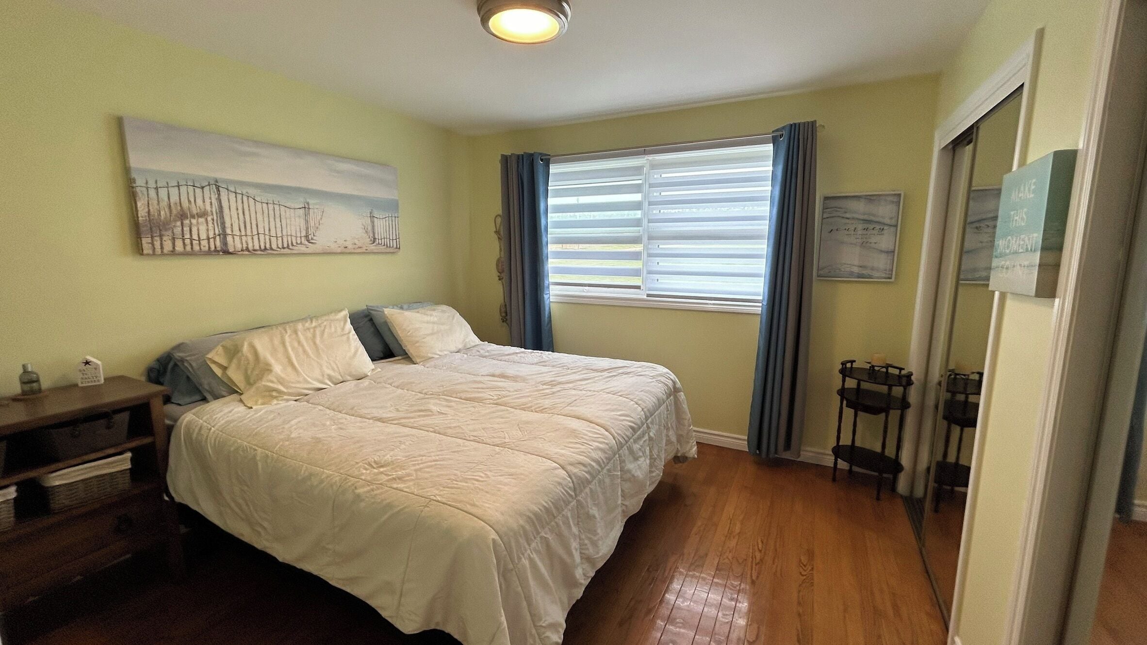 Une chambre à coucher avec un lit, une table de chevet, une fenêtre avec des stores, un tableau au mur et une porte avec une pancarte.