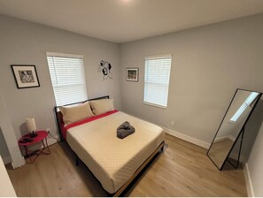 Ein Schlafzimmer mit einem Bett, zwei Fenstern mit Jalousien, einem Nachttisch mit Lampe und einem Spiegel an der Wand.