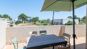 Eine Terrasse mit Tisch und Stühlen, einem grünen Sonnenschirm und Blick auf das Meer und Gebäude.