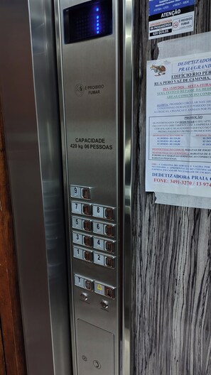 An elevator control panel with a digital display, keypad, and a sign with text in Portuguese. - Apartment for rent for short-term stays or weekends. (Praia Grande)