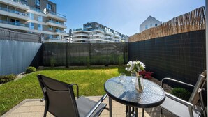 Een terras met een ronde glazen tafel, twee stoelen, een vaas met bloemen en een uitzicht op een meerhoog gebouw op de achtergrond.