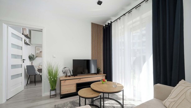 Een moderne woonkamer met een bank, twee bijzettafels, een televisie op een houten kast en een uitzicht op de stad door grote ramen.