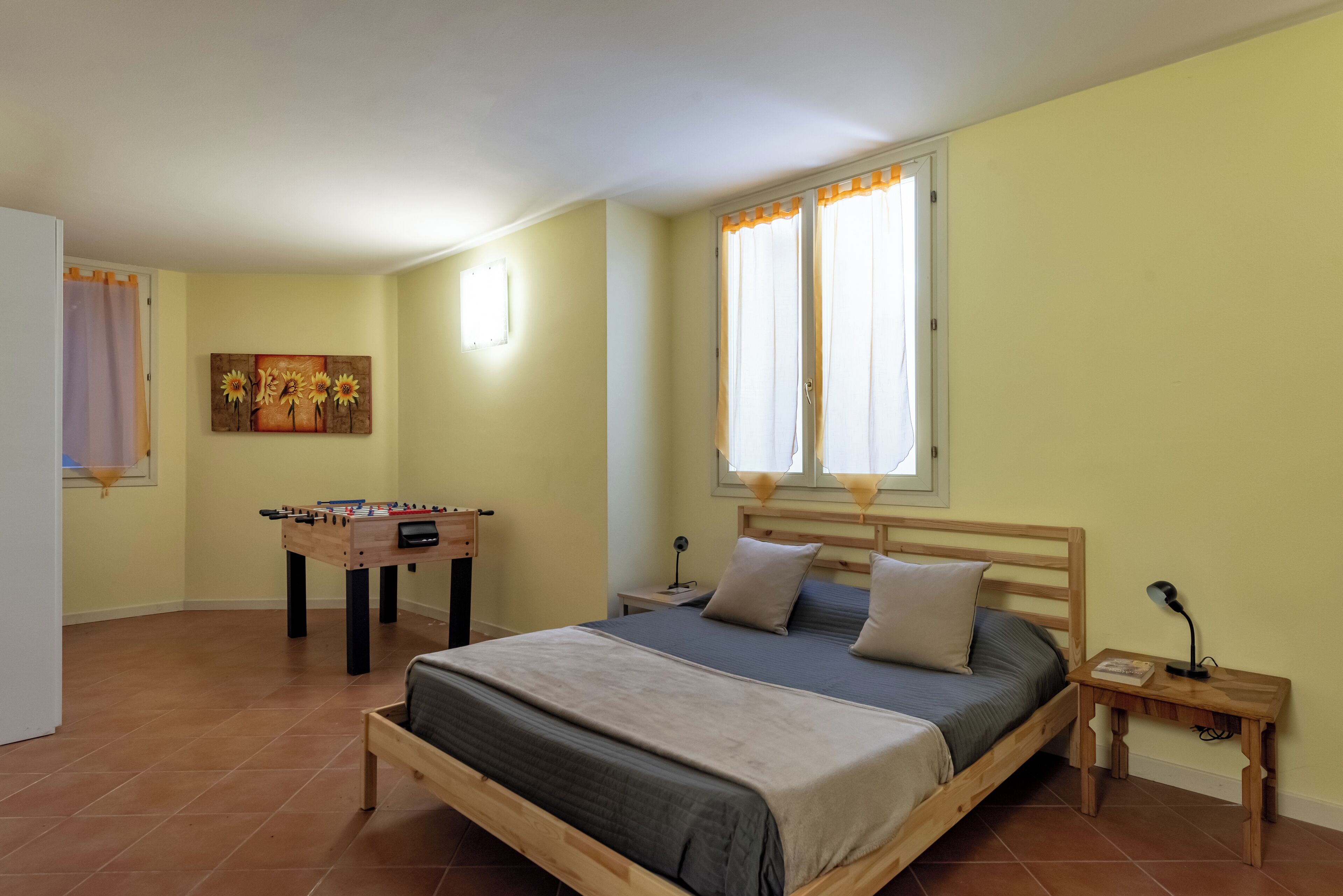 Une chambre à coucher avec un lit, une tête de lit en bois, une table de chevet, un bureau avec un baby-foot et une fenêtre avec des rideaux.