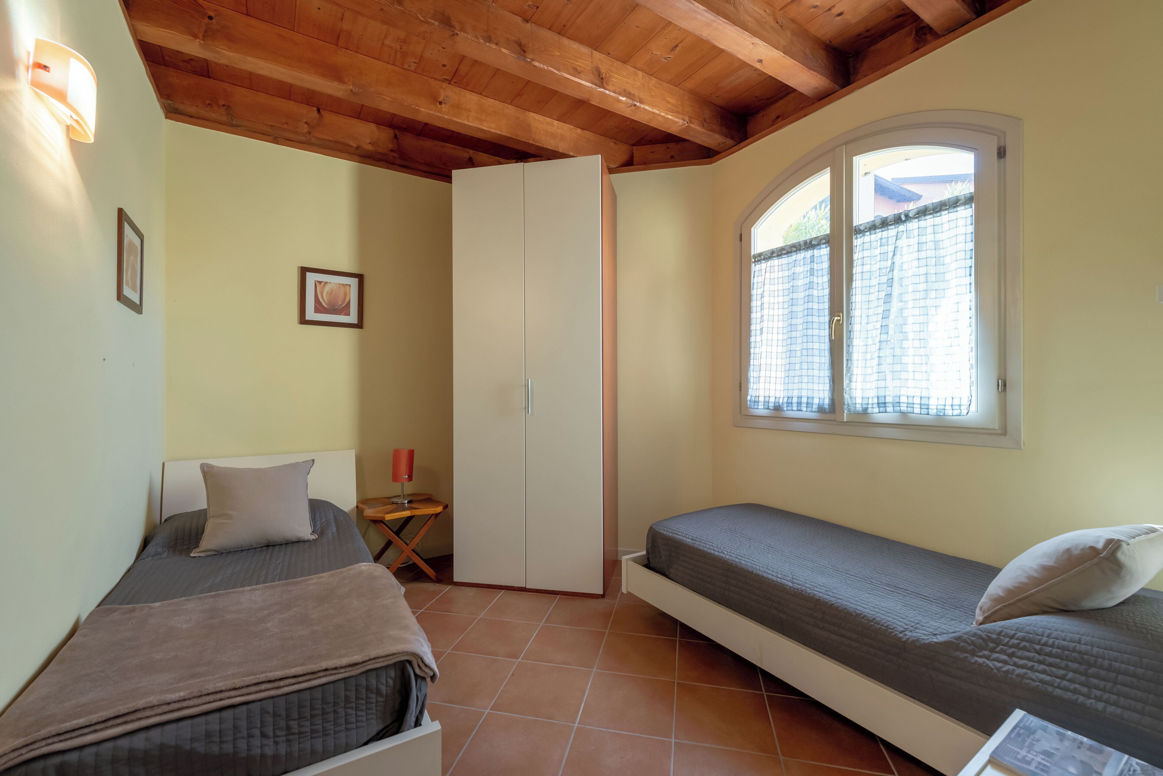 A small room with two beds, a wardrobe, a window with curtains, and a wall-mounted light fixture.