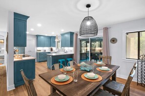 A modern kitchen with teal cabinetry, a central island, and a dining area with a wooden table set for four. - Charming remodeled 4th row House walk Beach, Coligny Plaza & Celebration Park! (Hilton Head Island)