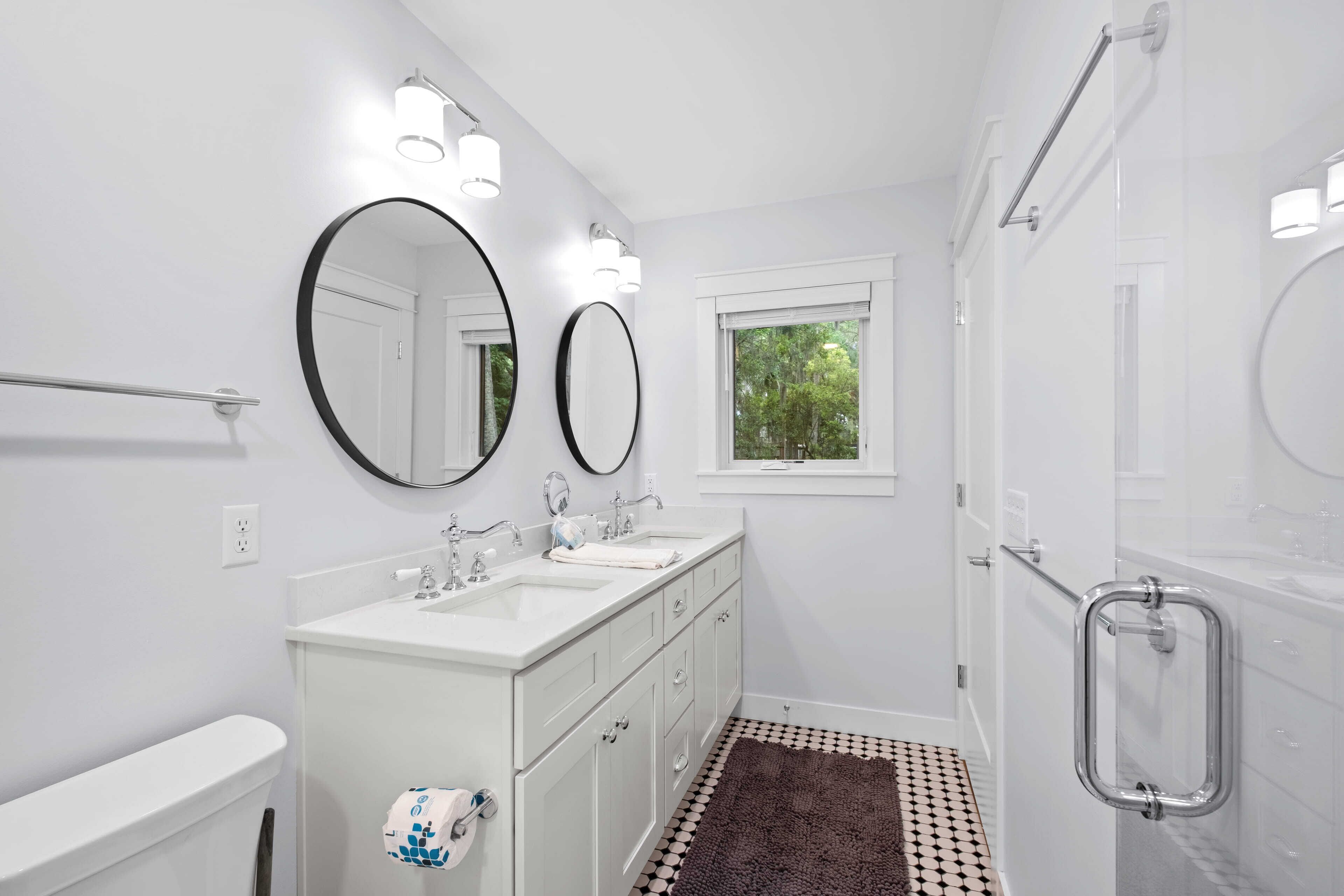 A modern bathroom with two sinks, a toilet, a shower with grab bars, and a window with a view of greenery.