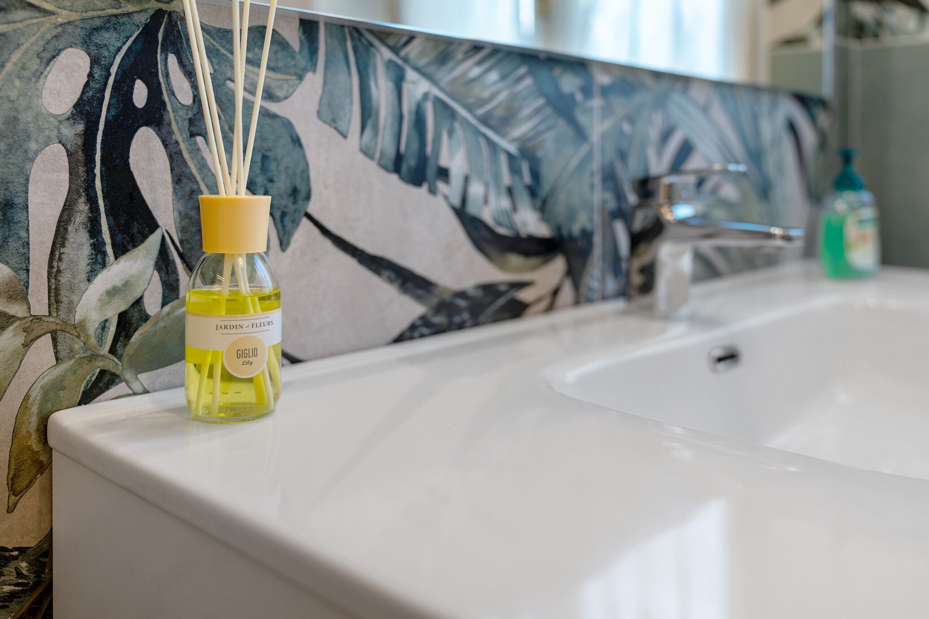 Un bagno con un lavandino bianco, uno schienale decorato con piastrelle e un flacone di profumo sul ripiano.