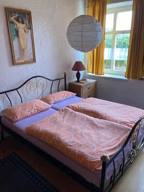 Ein Bett mit zwei Kissen, einer Nachttischlampe, einem Weidenbeistelltisch, einem gerahmten Bild an der Wand und einem Fenster mit Vorhängen.