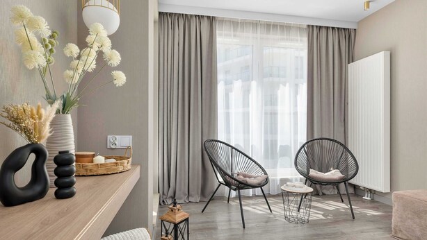 A modern living room with a wooden floor, two black chairs, a white sofa, a wooden side table, a vase with flowers, and a large window with curtains. - Modern Apartment with a Balcony | Pool, Spa, Gym (Międzyzdroje)