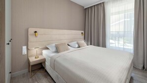 A hotel room with a large bed, bedside table with a lamp, a power outlet, and a window with curtains. - Modern Apartment with a Balcony | Pool, Spa, Gym (Międzyzdroje)