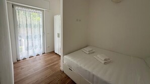 Ein Schlafzimmer mit Holzboden, einem Einzelbett mit weißer Bettwäsche, einem weißen Kleiderschrank und einer Glastür zum Außenbereich.