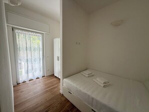 Ein Schlafzimmer mit Holzboden, einem Einzelbett mit weißer Bettwäsche, einem weißen Kleiderschrank und einer Glastür zum Außenbereich.