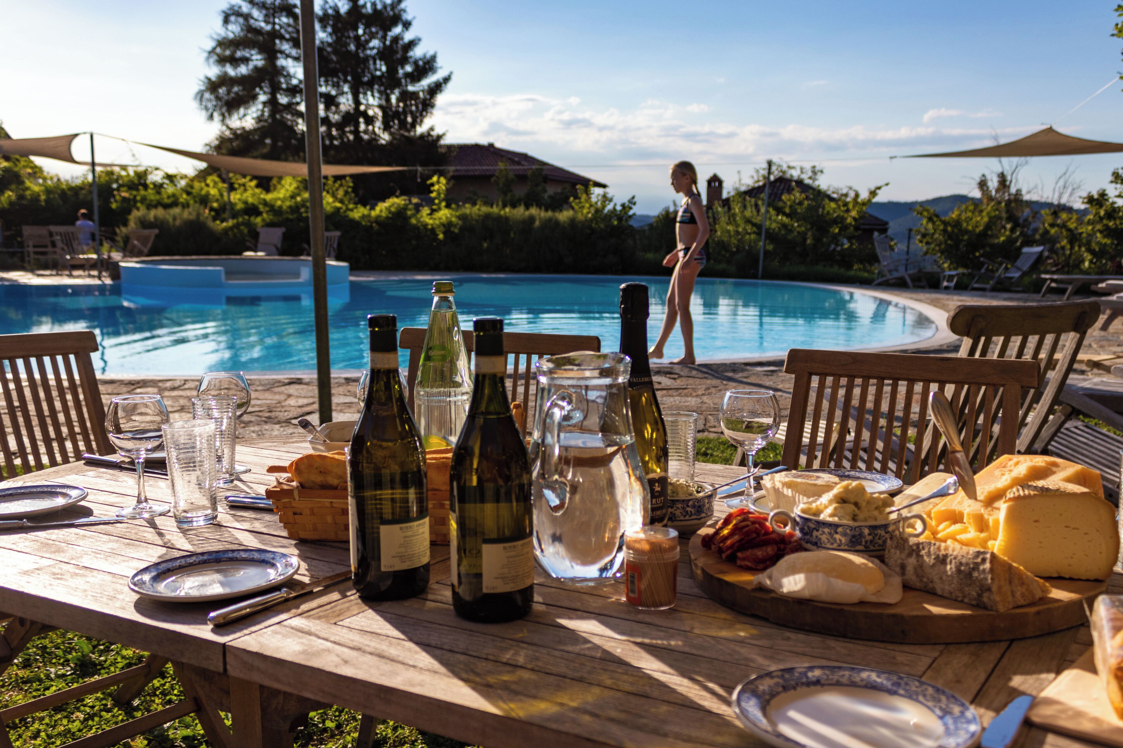 Ein Holztisch gedeckt mit Tellern, Weinflaschen, Gläsern und einer Käseplatte am Pool.
