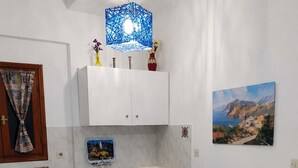 A compact kitchen with white cabinets, a small fridge, a glass dining table with white chairs, and a blue hanging light fixture. - Skopelos Cozy Escape Near The Forest the Sea (Skopelos)