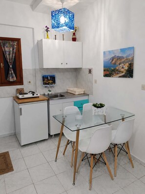 Eine kompakte Küche mit weißen Schränken, einem kleinen Kühlschrank, einem Glastisch mit weißen Stühlen und einer blauen Pendelleuchte.